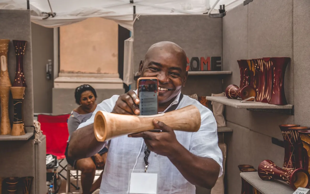 Un artisan souriant présente une pièce en bois qu'il vient de photographier avec son téléphone sur son stand de marché